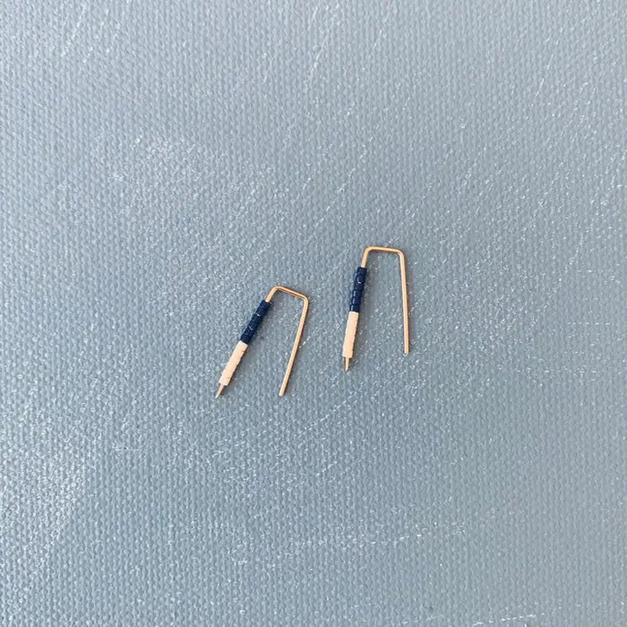 The ALICE RISE LITTLE QUILL EARRINGS IN NAVY AND CHALK are two small, L-shaped gold filled earrings with deep navy centers, shown parallel on a textured gray surface with their tips pointing downward.