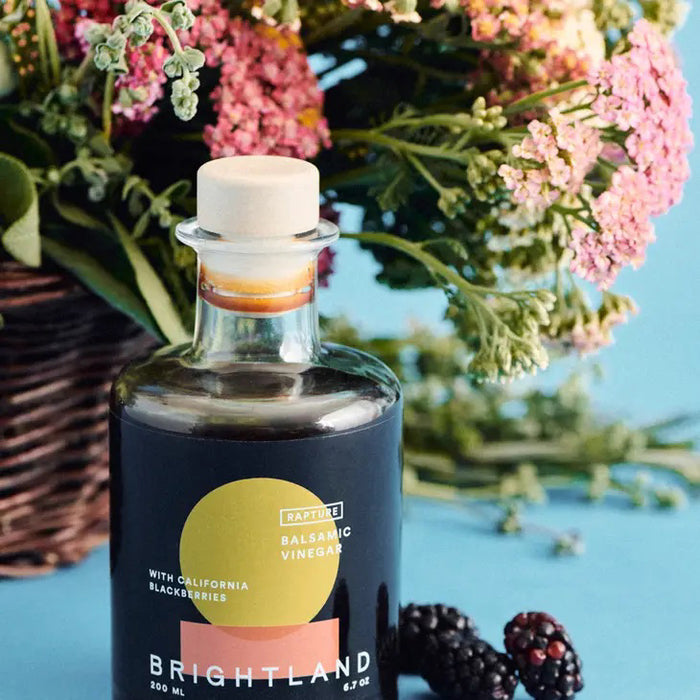 A clear glass bottle of BRIGHTLAND 'Rapture' Blackberry Balsamic Vinegar, 200 ml, featuring a red and yellow circle label stating “Balsamic Vinegar with California & triple crown blackberries.”.