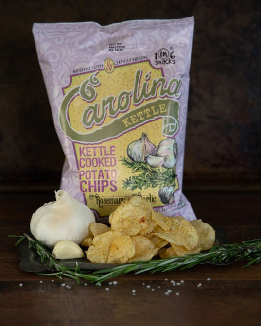 A pink 5oz bag of 1 IN 6 SNACKS CAROLINA KETTLE - ROSEMARY & GARLIC CHIPS features gourmet rosemary garlic flavor, illustrated with garlic and rosemary, plus non-GMO and gluten-free labels on the front.