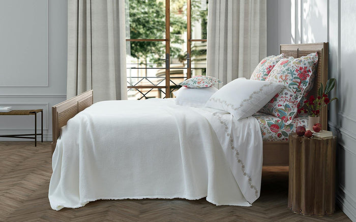 The Matouk Selah Queen Coverlet from the FEARRINGTON LIFESTYLE BEDDING COLLECTION is neatly folded with a matching pillow on a gray surface, set against a textured wall and green floral accents. Made of cotton in Portugal by Matouk.