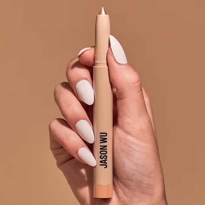 A hand with manicured nails holds a JEWEL STICK EYESHADOW - SPARKLE PEARL by JASON WU BEAUTY, against a light brown background.