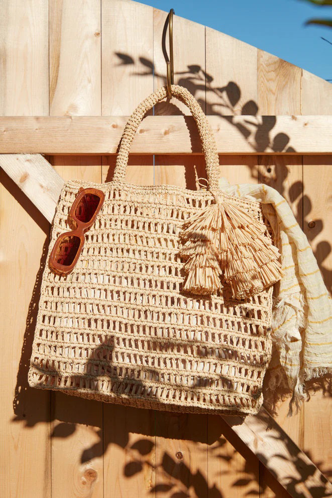 The MAR Y SOL - GEORGIA TOTE by MAR Y SOL is a rectangular crocheted raffia bag with two handles and a decorative tassel, shown against a white background.
