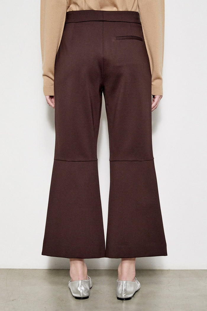 A person models the MEIMEIJ Crop Ponte Pant with Belt, shown in brown with pockets, paired with a beige top and silver flats. Only the lower half is visible against a plain white wall and concrete floor.