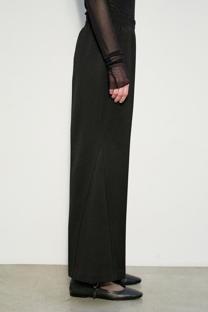 A person stands against a white wall wearing MEIMEIJ - STRIGHT LEG PULL ON PONTE PANT by MEIMEIJ with a sheer black long-sleeve top and black shoes. Only the lower body is visible; the upper body and head are not shown.