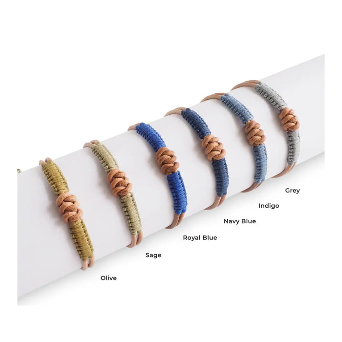 The MAKARLA MEN'S KNOT LEATHER BRACELET ADJUSTABLE IN NAVY is displayed on a white cylindrical stand, labeled by color. This handmade bracelet adds unique style and can be easily adjusted for the perfect fit.