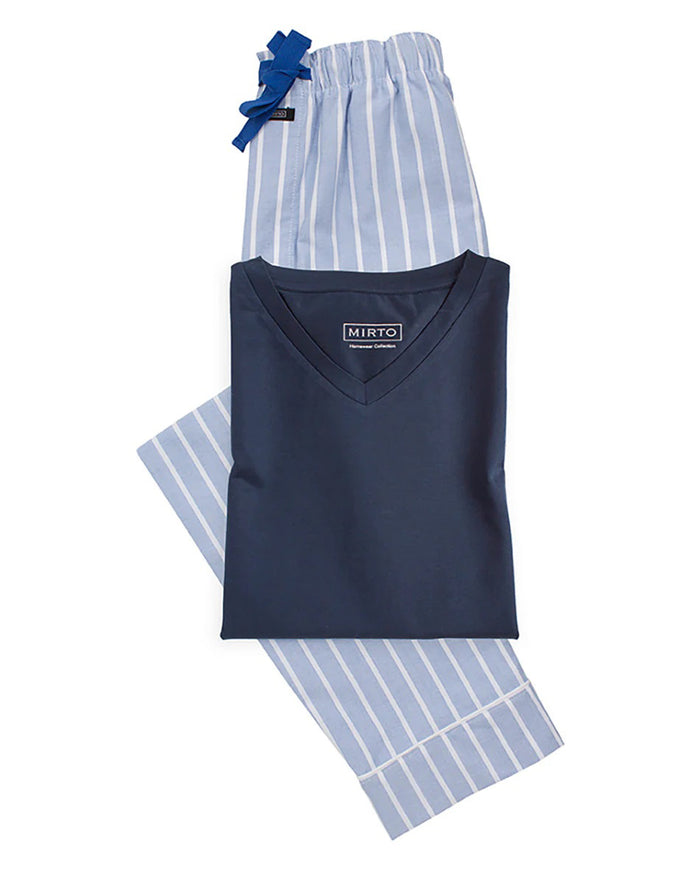 A man stands barefoot on a plain background, facing away, wearing the MIRTO 1956 - OXFORD PAJAMA SET: a dark blue top paired with loose, light blue cotton pants featuring white vertical stripes by MIRTO 1956.