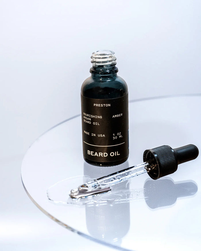 A small dark glass bottle of PRESTON - BEARD OIL by PRESTON, with a dropper cap, sits on a light surface casting a soft shadow. The label displays "PRESTON - BEARD OIL" along with product details in white text.