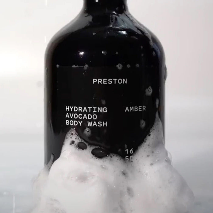 A black bottle labeled "Preston Hydrating Avocado Body Wash Amber" sits upright with soapy foam covering its base against a white background.