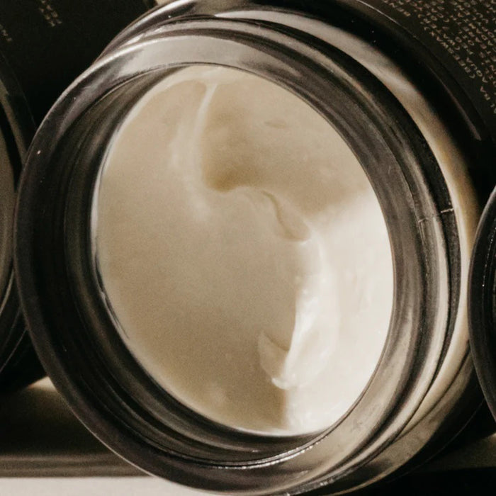 A black jar labeled "PRESTON - CREAM POMADE UNSCENTED" rests on a beige surface with a soft shadow, highlighting its matte finish and light hold formula by PRESTON, set against a neutral background.
