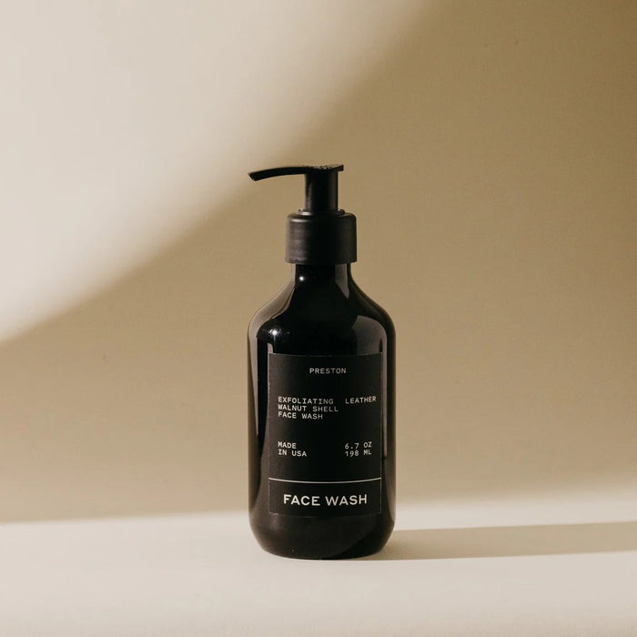 A black pump bottle labeled "PRESTON - FACE WASH" by PRESTON sits on a beige surface in soft, natural light. The minimalist label notes it’s a resurfacing exfoliating cleanser with walnut shells to unclog pores, made in the USA.