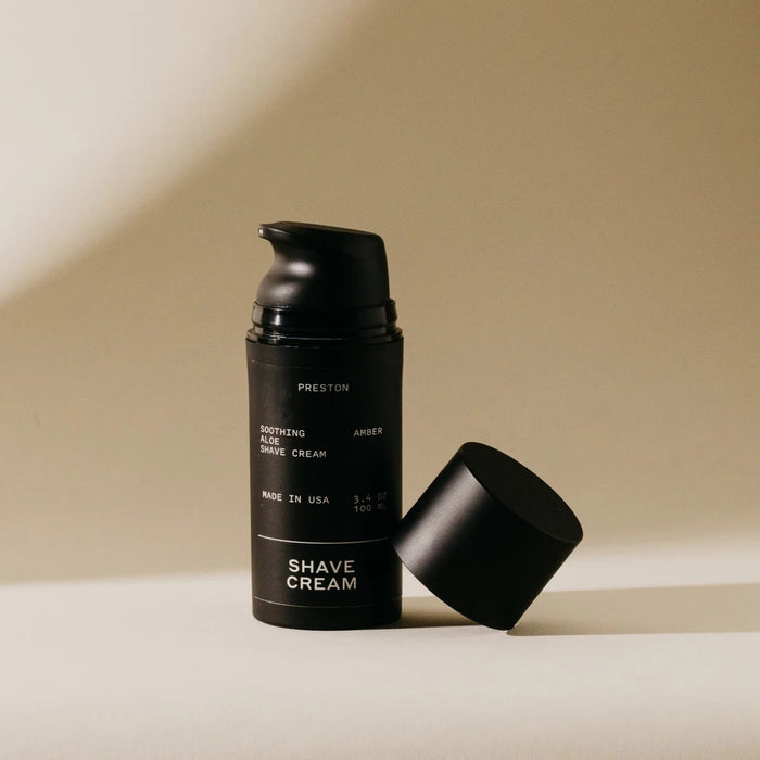 A black cylindrical bottle of PRESTON - SHAVE CREAM stands on a light beige surface with a soft shadow, featuring minimalist packaging with white text that highlights its cooling formula infused with soothing aloe vera.