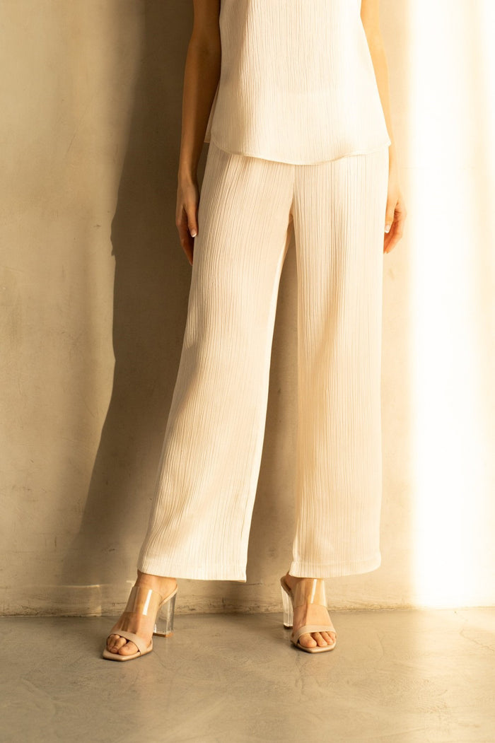 A woman smiles against a white backdrop, wearing a black top and ROSE AND CROWN - SATIN RIBBED PANT. She accessorizes with layered necklaces and a large black handbag, posing with one leg slightly raised for a relaxed look.