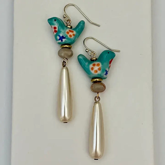 The SANDRINE FRANCE STUDIO GEMMA Earrings feature handmade turquoise ceramic birds with painted details and elongated pearl-like drops, crafted in NYC. Shown on a clear stand against a plain, neutral background.