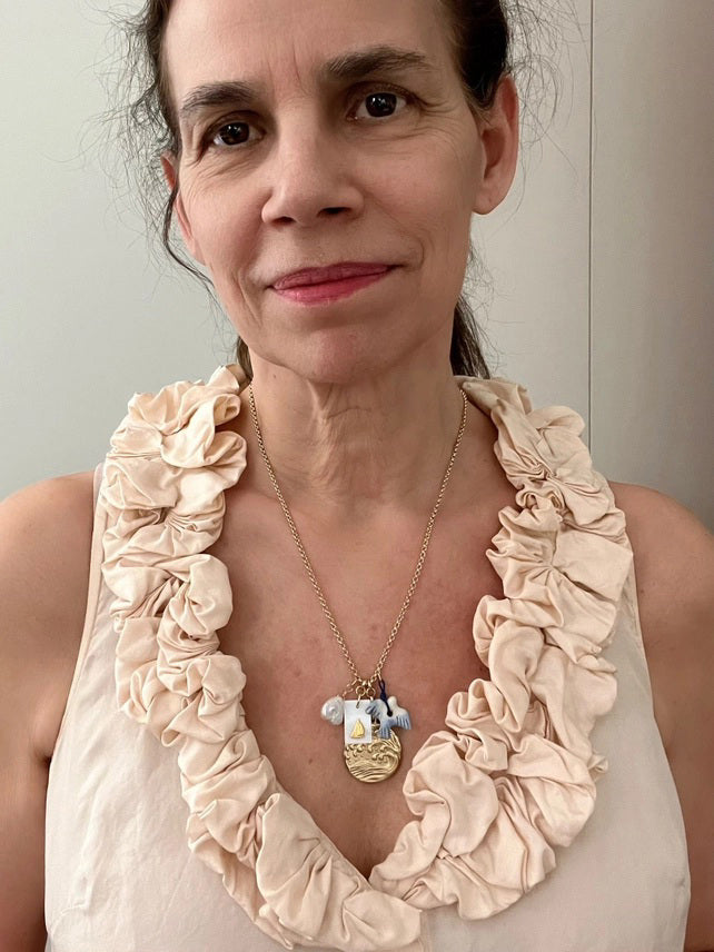 The SANDRINE FRANCE STUDIO WREN NECKLACE is a handmade gold necklace from NYC featuring four charms—a white shell, a rectangular gold sailboat tag, a gold wave pendant, and a blue-white fish—on a white jewelry stand.