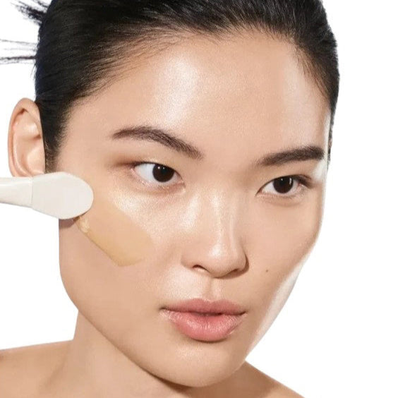 A woman with radiant skin applies liquid foundation to her cheek, showing off her clear, luminous complexion—thanks to U BEAUTY RESURFACING FLASH PEEL, which gently exfoliates and brightens for an even base.