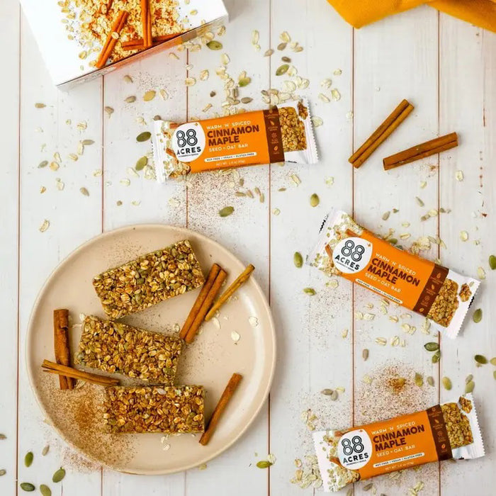 Image of an 88 ACRES Cinnamon Maple Seed + Oat Bar. The packaging is white with an orange section that displays the brand "88 ACRES" and the flavor details. The package showcases the nut-free, vegan, and gluten-free seed bar sprinkled with oats and seeds.