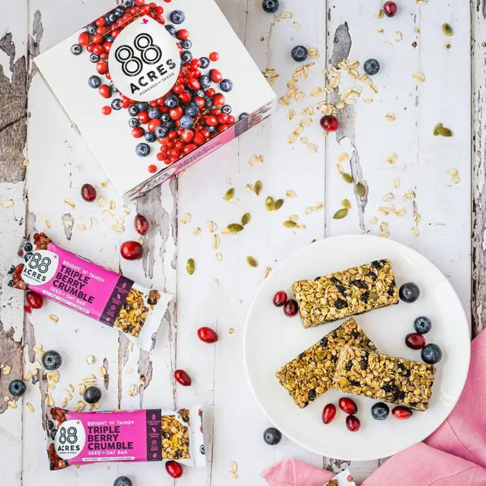 An 88 ACRES - TRIPPLE BERRY CRUMBLE bar by 88 ACRES, a nut-free vegan oat bar, rests on a light wooden surface in its white and purple berry-themed wrapper featuring nut-free, vegan, and gluten-free labels.