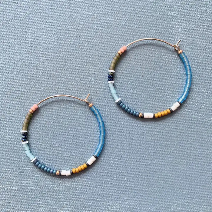 The ALICE RISE - BIG COLORLOOP EARRINGS IN SPRINGWATER by ALICE RISE feature two large hoops adorned with small, colorful Japanese glass beads, elegantly arranged and displayed on a textured light grey surface.