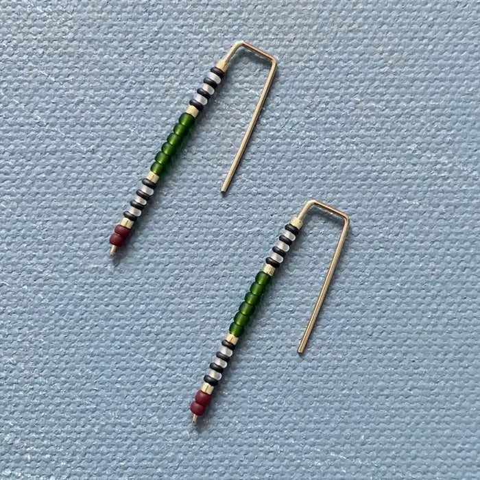 The ALICE RISE - BIG QUILL EARRINGS IN FOREST GREEN by ALICE RISE feature 14k gold-filled hooks and small red, green, black, and white beads. Displayed parallel on a textured light blue background, their hooks face opposite directions.
