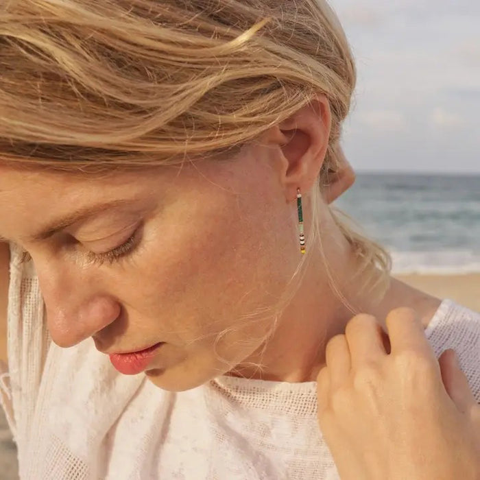 The ALICE RISE - BIG QUILL EARRINGS IN FOREST GREEN by ALICE RISE feature 14k gold-filled hooks and small red, green, black, and white beads. Displayed parallel on a textured light blue background, their hooks face opposite directions.
