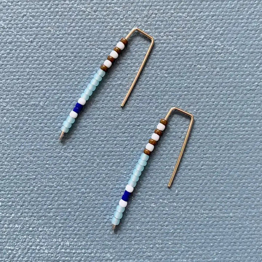 The ALICE RISE - BIG QUILL EARRINGS IN SKY BLUE feature gold wire and vertical rows of small beads in light blue, white, brown, and dark blue. Handmade by ALICE RISE, these earrings are perfect for sensitive skin and displayed on a textured light gray surface.