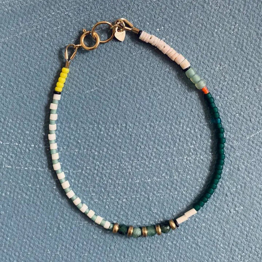 The ALICE RISE - EMERALD BRACELET by ALICE RISE showcases handmade North Carolina artistry with white, yellow, green, teal, and metallic gold beads and a gold clasp, elegantly displayed on a textured blue surface.