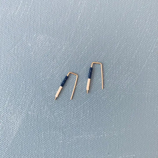 The ALICE RISE LITTLE QUILL EARRINGS IN NAVY AND CHALK are two small, L-shaped gold filled earrings with deep navy centers, shown parallel on a textured gray surface with their tips pointing downward.