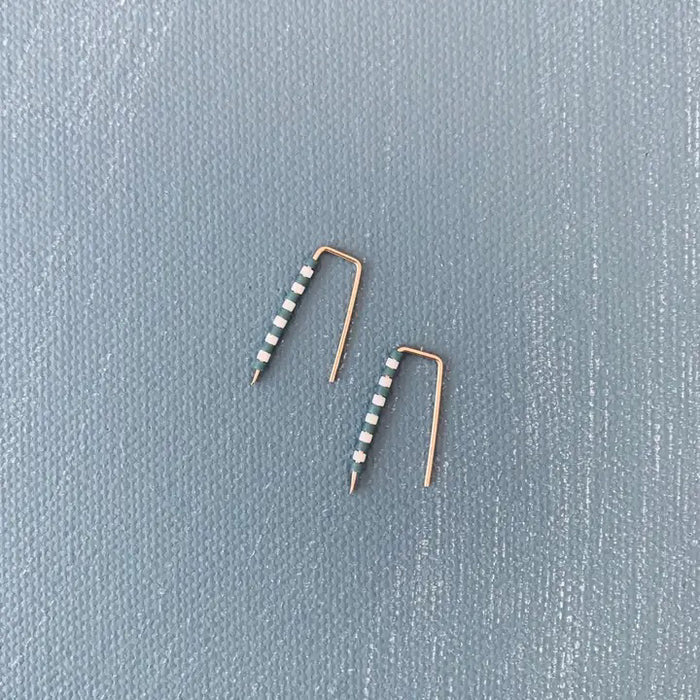 The ALICE RISE - LITTLE QUILL EARRINGS IN SLATE BLUE STRIPE feature unique 14k gold filled posts adorned with alternating white and green beads, arranged parallel on a textured light gray surface.