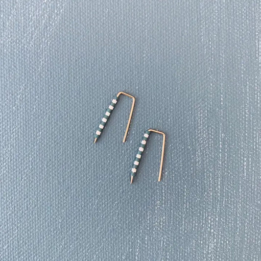 The ALICE RISE - LITTLE QUILL EARRINGS IN SLATE BLUE STRIPE feature unique 14k gold filled posts adorned with alternating white and green beads, arranged parallel on a textured light gray surface.