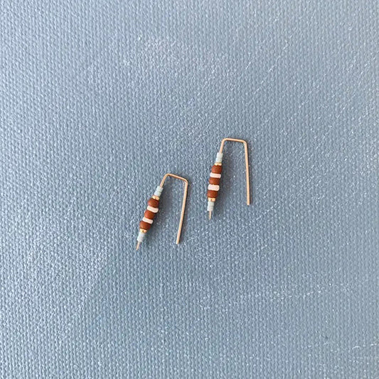 ALICE RISE - LITTLE QUILL EARRINGS IN TERRACOTTA by ALICE RISE rest on a textured gray surface, their unique design reminiscent of bent electronic resistors—perfect inspiration for handmade jewelry enthusiasts.
