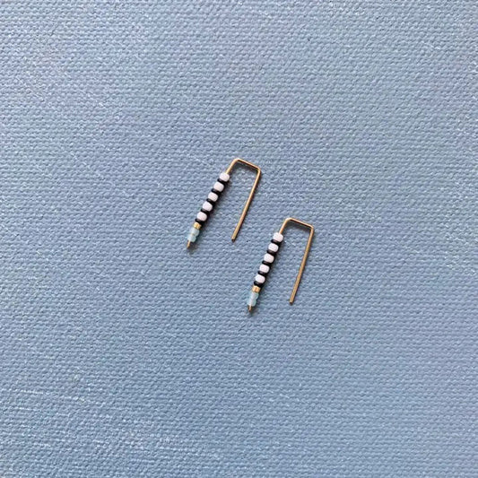 The ALICE RISE - LITTLE QUILL EARRINGS IN TURQUOISE by ALICE RISE, featuring minimalist 14k gold filled design and turquoise beads, are displayed on a textured light blue background—perfect for handmade jewelry lovers.