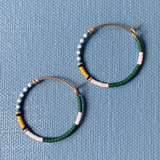 The ALICE RISE MIDI COLORLOOP EARRINGS IN FAIRWAY GREEN feature round 14k gold filled hoops with green, white, yellow, and black Japanese glass beads, displayed on a textured light blue background.
