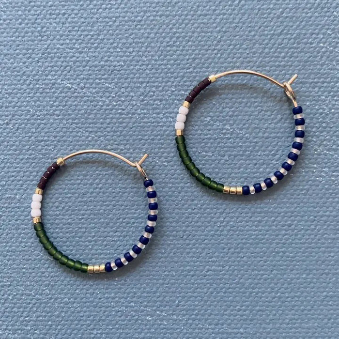 The ALICE RISE - MIDI COLORLOOP EARRINGS IN OLIVE by ALICE RISE feature 14k gold filled hoops adorned with white, navy blue, green, burgundy, and gold Japanese glass beads, displayed on a textured light blue surface.