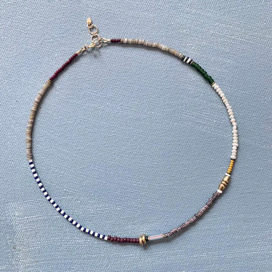 The ALICE RISE - PEARL MIXED MEDIA NECKLACE by ALICE RISE features small, multicolored vintage glass beads arranged in sections and finished with a 14k gold-filled clasp, displayed in a circle on a textured light blue surface.