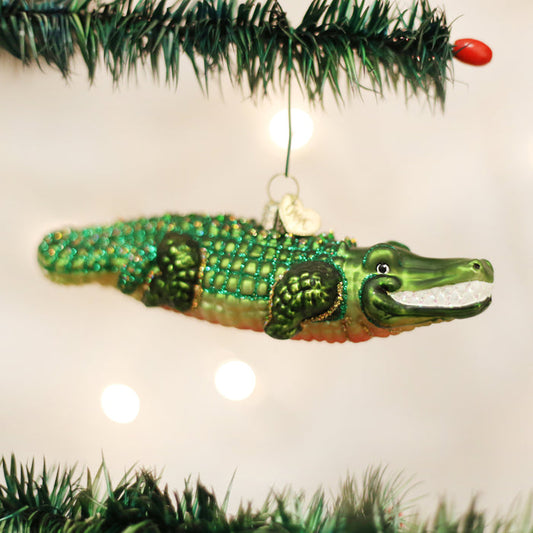 A shiny green alligator ornament hangs from a Christmas tree branch, surrounded by green needles and small white lights.