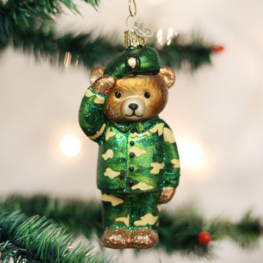 A Christmas tree ornament shaped like a teddy bear in a glittery green camouflage uniform and beret, saluting while hanging from a tree branch.