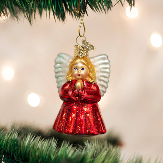 A glass angel ornament with white wings and a glittery red dress hangs from a green Christmas tree branch, with soft white lights blurred in the background.