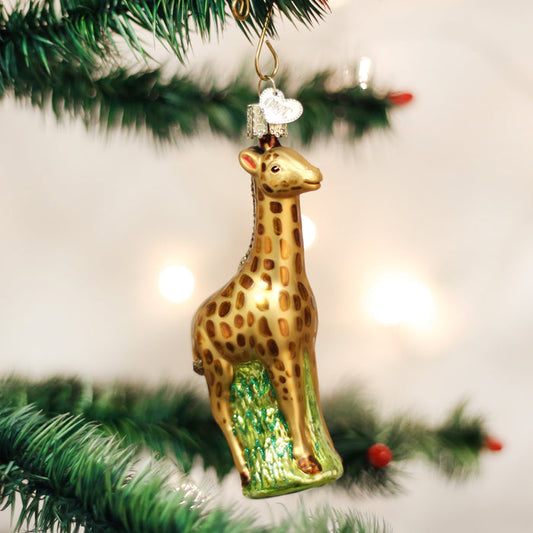 A glass ornament shaped like a giraffe with brown spots hangs from a green Christmas tree branch, with blurred white lights in the background.