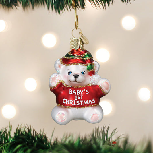 A Christmas tree ornament shaped like a white teddy bear wearing a red outfit with BABYS 1ST CHRISTMAS written on it and a red-and-green hat, hanging on a tree with blurred lights in the background.