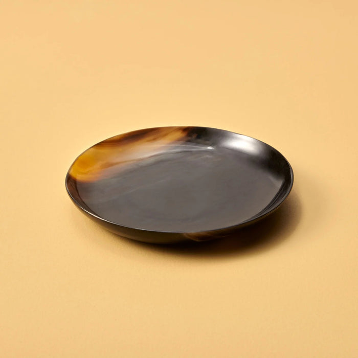The BE HOME - SPLATTER HORN PLATE SMALL by BE HOME, INC is a round, shallow plate handmade in Vietnam from water buffalo horn, showcasing a dark brown and amber marbled pattern on a beige surface.