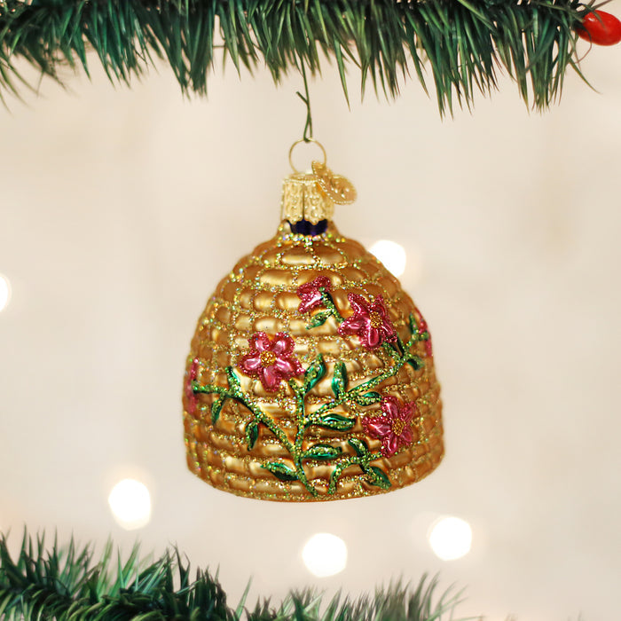 A gold, beehive-shaped Christmas ornament decorated with pink flowers and green leaves hangs from a green branch, with soft white lights glowing in the blurred background.