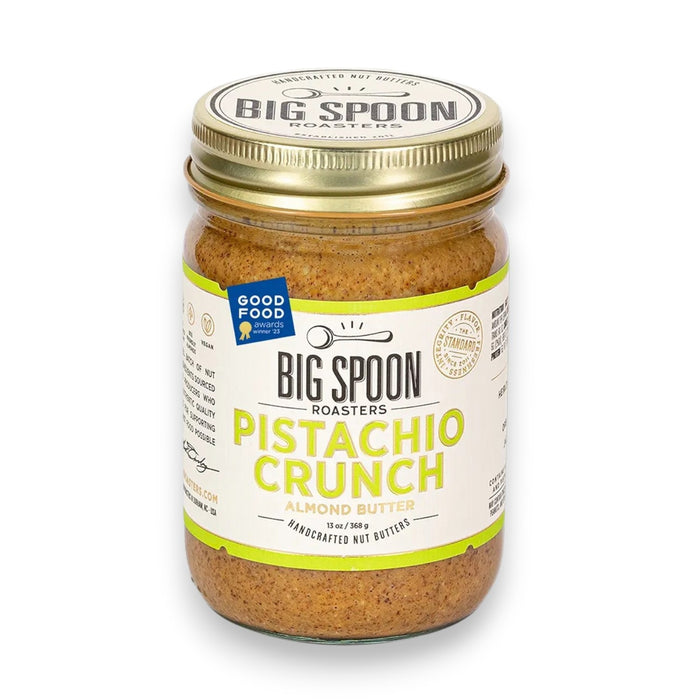 A clear glass jar of BIG SPOON ROASTERS - PISTACHIO & ALMOND CRUNCH NUT BUTTER, sealed with a gold lid and a white label with green and yellow text. The nut butter is made from heirloom mission almonds by BIG SPOON ROASTERS.
