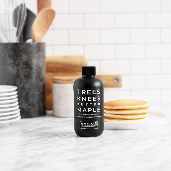A black bottle labeled "Bushwick Kitchen - Trees Knees Butter Maple Syrup" features white text, "Born in Brooklyn, NY," vegan butter flavor highlights, and displays "Net Wt 11.5 oz (326g).