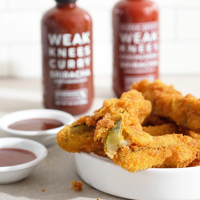 A 10.5 oz (298g) bottle of BUSHWICK KITCHEN 'WEAK KNEES' CURRY SRIRACHA SAUCE with a black twist cap, white front text, and a Gochujang chile paste blend for a unique flavor kick.