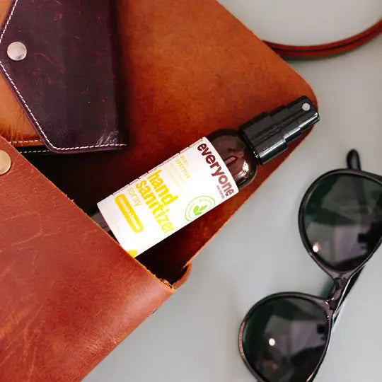 A 2 oz bottle of EVERYONE FOR EVERYBODY Coconut & Lemon Hand Sanitizer Spray is partially visible inside an open brown leather bag, beside black sunglasses and a dark wallet on a light surface.