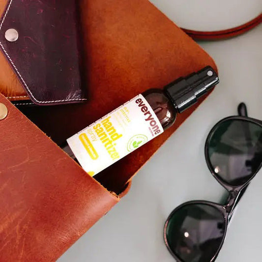 A 2 oz bottle of EVERYONE FOR EVERYBODY Coconut & Lemon Hand Sanitizer Spray is partially visible inside an open brown leather bag, beside black sunglasses and a dark wallet on a light surface.