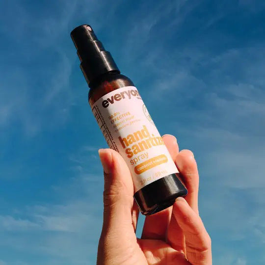 A 2 oz bottle of EVERYONE FOR EVERYBODY Coconut & Lemon Hand Sanitizer Spray is partially visible inside an open brown leather bag, beside black sunglasses and a dark wallet on a light surface.