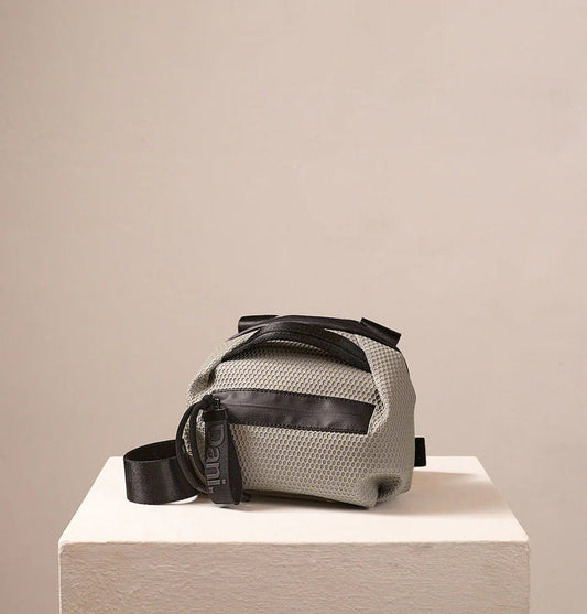 The DANIELLA LEHAVI BONNIE MINI TRAVEL CROSSBODY, a small brown vegan bag with black accents and strap, is displayed on a white pedestal against a plain beige background.