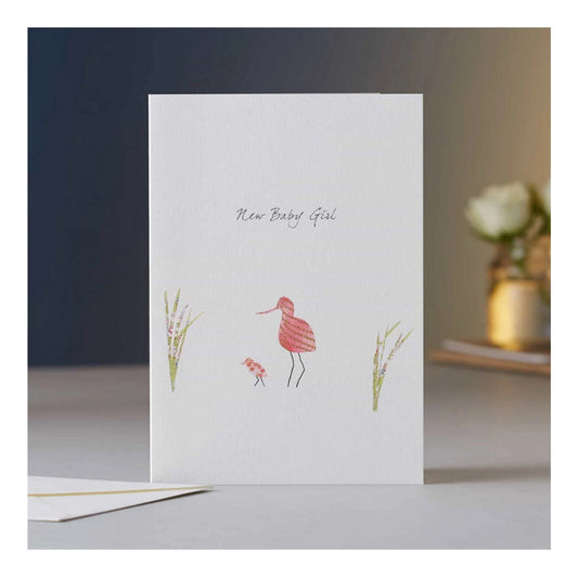The ELOISE HALL - AVOCET BABY GIRL CARD by ELOISE HALL LTD features "New Baby Girl" above pink bird illustrations—one large, one small—amid grass on a white background. The luxury card stands upright on a table.
