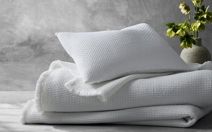 The Matouk Selah Queen Coverlet from the FEARRINGTON LIFESTYLE BEDDING COLLECTION is neatly folded with a matching pillow on a gray surface, set against a textured wall and green floral accents. Made of cotton in Portugal by Matouk.